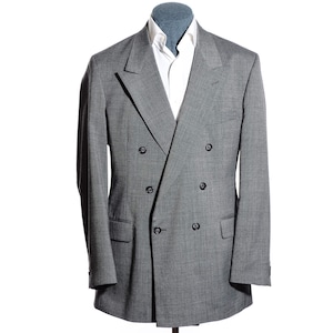 May include: A double-breasted gray blazer featuring a subtle plaid pattern, wide peak lapels, and six dark buttons. The blazer is worn over a crisp white collared shirt, showcasing a structured silhouette with long sleeves and two flap pockets.