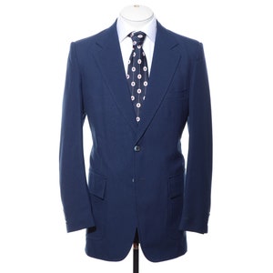 May include: A navy blue blazer with a single-button closure, paired with a white dress shirt and a patterned tie. The blazer has two front pockets and is displayed on a mannequin. The tie features a repeating pattern of white circles on a dark background.