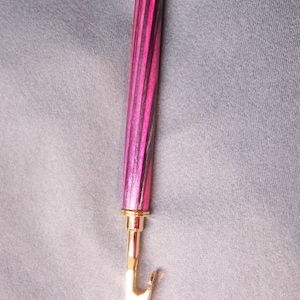 May include: A black and pink wooden pen with a gold clip and a gold top. The pen is on a grey background.