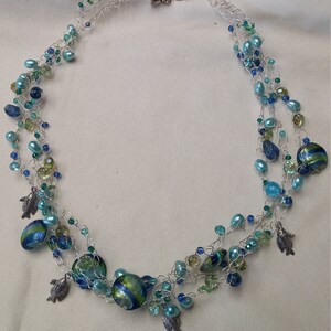 Puede incluir: Un collar de múltiples hilos con cadena plateada y cuentas de vidrio azul y verde. El collar presenta pequeños colgantes de peces plateados.