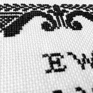 May include: A white fabric featuring black cross-stitch embroidery. The design includes a decorative border with intricate patterns and the text "EW," stitched in a bold, dark font. The needlework displays a clear, textured grid pattern.