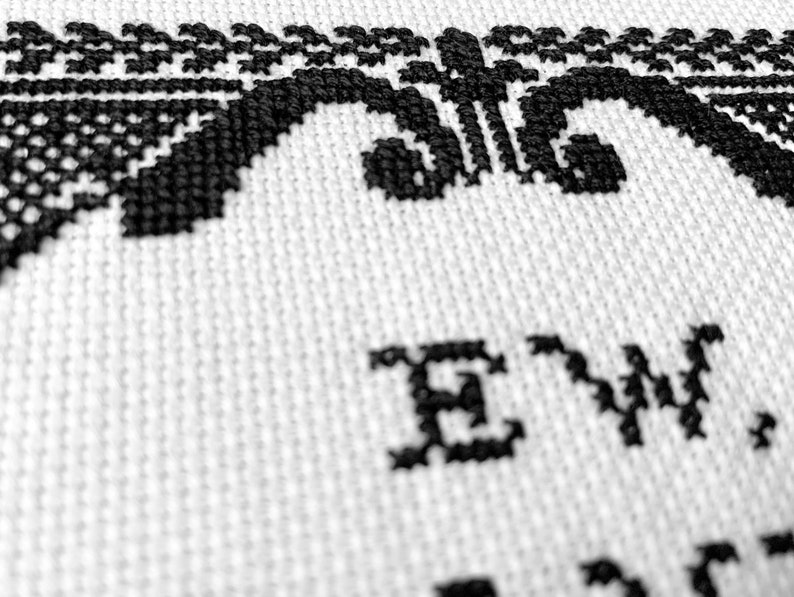 May include: A white fabric featuring black cross-stitch embroidery. The design includes a decorative border with intricate patterns and the text "EW," stitched in a bold, dark font. The needlework displays a clear, textured grid pattern.
