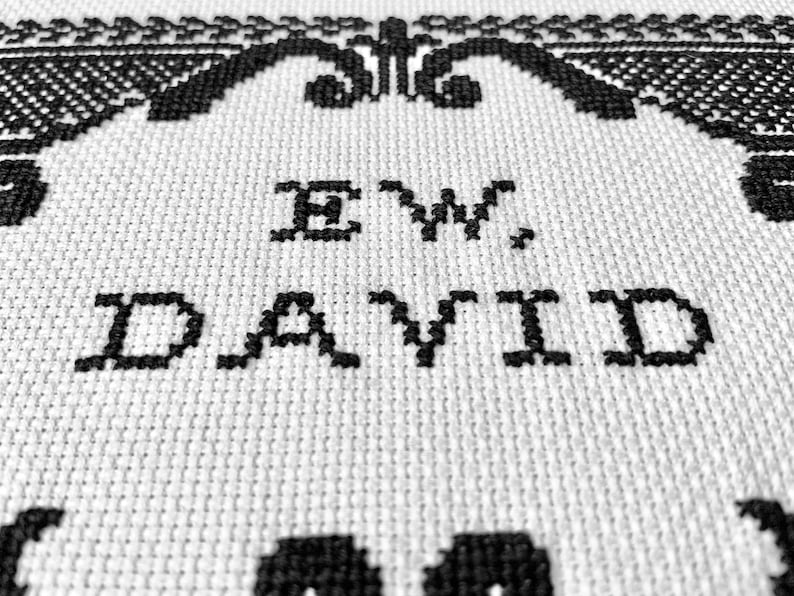 May include: A white fabric piece featuring black cross-stitch embroidery. The text "EW, DAVID" is centered in the middle, surrounded by a decorative black border pattern. The fabric has a visible grid-like texture characteristic of Aida cloth.