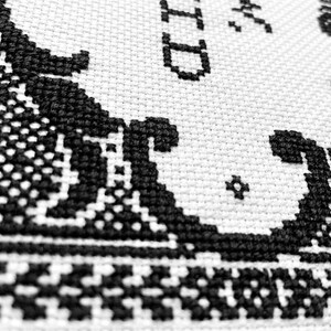 May include: A white fabric with black cross-stitch embroidery featuring intricate scrollwork patterns and partial text that includes the letters ID. The needlework displays a dense, textured grid of stitches forming decorative borders.
