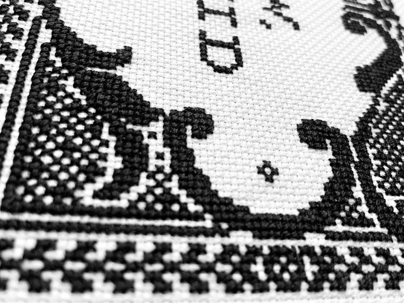 May include: A white fabric with black cross-stitch embroidery featuring intricate scrollwork patterns and partial text that includes the letters ID. The needlework displays a dense, textured grid of stitches forming decorative borders.