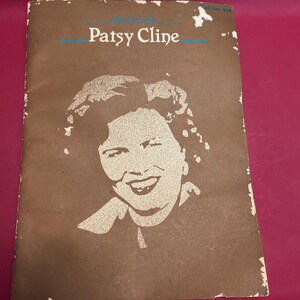 Könnte beinhalten: Ein braunes Liederbuch-Cover aus Papier mit einem weißen grafischen Porträt von Patsy Cline. Der Text oben lautet The Best of Patsy Cline in weißer Schrift mit blauen horizontalen Linien. Die Ränder des Covers weisen Abnutzungserscheinungen auf.