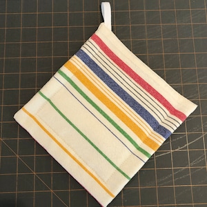 Può includere: Presina color crema con strisce colorate rosse, blu, gialle e verdi. Le strisce diagonali attraversano la forma quadrata. Un anello bianco è attaccato per appendere. La presina è in tessuto e ha un bordo cucito.
