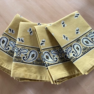 May include: Four yellow fabric napkins with a black and white paisley pattern border. The napkins are folded and stacked on top of each other.