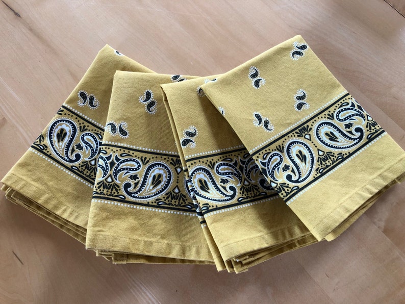 May include: Four yellow fabric napkins with a black and white paisley pattern border. The napkins are folded and stacked on top of each other.