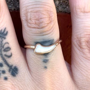 May include: A gold-toned ring featuring a curved, horn-shaped inlay made of a smooth, white, pearlescent material. The band is thin and polished, with the central decorative element positioned horizontally across the finger.