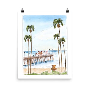 May include: Watercolor print of a pier with palm trees, a lifeguard tower, and a beach. The pier has a building with red umbrellas. The sky is blue with a few birds. The artwork is signed by the artist.