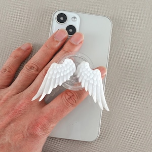 May include: White angel wing phone grip attached to a silver smartphone. The wings are detailed with individual feathers. The grip is clear and circular, designed to adhere to the phone's surface for easy handling.