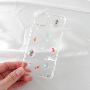 May include: A clear phone case featuring a design of red goldfish and blue bubbles. The case is made from a flexible, transparent material.