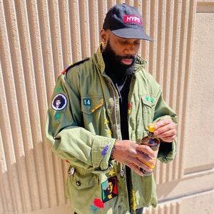 May include: A man wearing a green military jacket with various patches and a black baseball cap with the word "HYPE" in red letters. He is holding a glass bottle with a yellow cap.