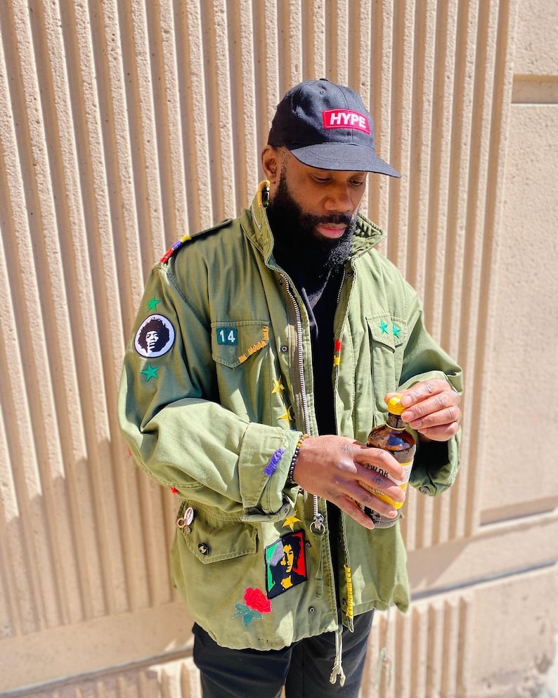May include: A man wearing a green military jacket with various patches and a black baseball cap with the word "HYPE" in red letters. He is holding a glass bottle with a yellow cap.