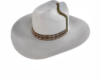 Könnte beinhalten: Ein weißer Cowboyhut mit breiter Krempe und einem dekorativen Band um den Kronenbereich. Das Band weist ein Muster aus kleinen, goldfarbenen Perlen auf. Ein vertikaler olivgrüner Streifen verläuft die Krone hinunter.