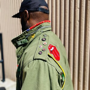 May include: A green military-style jacket with various patches, including a red tongue logo, a yellow star, and a red cross. The jacket has a zipper closure and a black button on the shoulder.