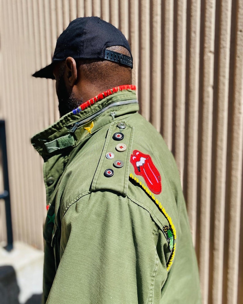 May include: A green military-style jacket with various patches, including a red tongue logo, a yellow star, and a red cross. The jacket has a zipper closure and a black button on the shoulder.