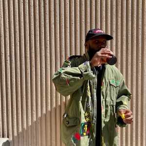 May include: A green military-style jacket with various patches and embroidery, including a portrait of Bob Marley, stars, and the word "Hype" on a black baseball cap. The jacket has a zipper closure and multiple pockets.