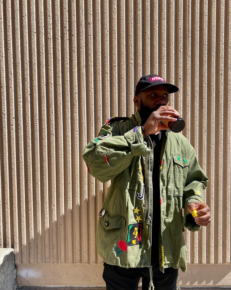May include: A green military-style jacket with various patches and embroidery, including a portrait of Bob Marley, stars, and the word "Hype" on a black baseball cap. The jacket has a zipper closure and multiple pockets.