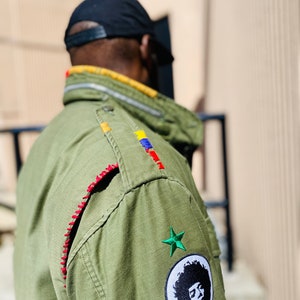 May include: A green military-style jacket with red stitching, colorful embroidery, and a patch with a black and white portrait of a person with an afro. The jacket also has two green star patches.