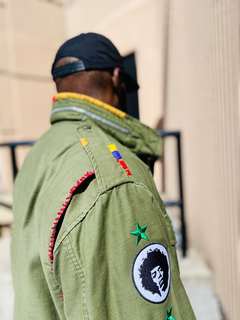 May include: A green military-style jacket with red stitching, colorful embroidery, and a patch with a black and white portrait of a person with an afro. The jacket also has two green star patches.