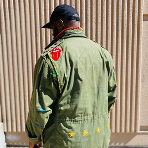 May include: A green military-style jacket with various patches, including a red tongue logo, a green leaf, and three yellow stars. The jacket has a black zipper and a black collar.