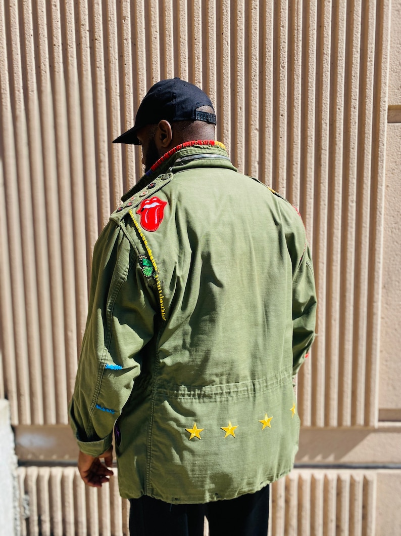 May include: A green military-style jacket with various patches, including a red tongue logo, a green leaf, and three yellow stars. The jacket has a black zipper and a black collar.