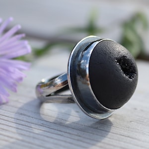 Puede incluir: Un anillo de plata con una piedra redonda negra engastada en un engaste de plata. La piedra tiene un pequeño agujero en el centro.