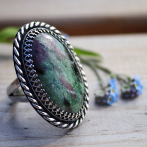 May include: A silver ring with a green and pink oval gemstone set in a twisted rope design band.