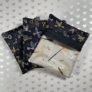 Peut inclure: Trois pochettes zippées en tissu noir avec des accents dorés. Les pochettes présentent un motif répété de clés, d'ailes et d'étoiles. La pochette à l'avant est doublée d'un tissu blanc avec un motif de baguette dorée et d'étoiles. Le texte "wing" et "mora" est visible sur la pochette.