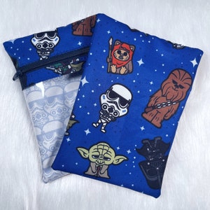 Könnte beinhalten: Eine blaue Reißverschlusstasche mit einem transparenten Plastikfenster. Die Tasche ist mit einem sich wiederholenden Muster von Star Wars-Charakteren verziert, darunter Yoda, Darth Vader, Chewbacca und Stormtrooper. Die Figuren sind im Cartoon-Stil gehalten und vor einem blauen Hintergrund mit weißen Sternen platziert.