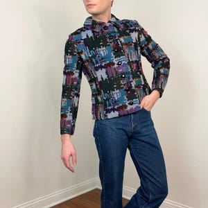 Può includere: Giacca fantasia con bottoni sul davanti nei toni del blu, viola e verde. La giacca ha un colletto e maniche lunghe. La persona nella foto indossa jeans blu.