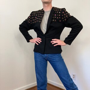 May include: A black blazer with a fitted silhouette and a single button closure. The blazer features a unique design with rhinestones on the shoulders. The model is wearing blue jeans.