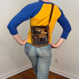 May include: A patchwork crossbody bag crafted from various shades of brown, tan, and navy blue suede leather. The bag features a front flap with a silver snap closure, a smaller front pocket with a snap, and a vertical side zipper.