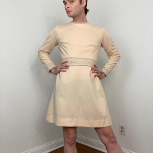 Könnte beinhalten: Ein cremefarbenes, langärmliges Kleid mit rundem Ausschnitt und ausgestelltem Rock. Das Kleid hat ein dekoratives Band an der Taille und Besätze an den Manschetten. Das Kleid ist knielang und hat eine Vintage-Ästhetik.