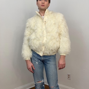 May include: Cream-colored faux fur jacket with a button-up front and a stand-up collar. The jacket has long sleeves and is paired with light blue, distressed jeans. The outfit is a vintage style.