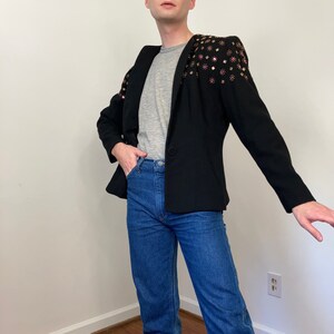 May include: A black blazer with a gray t-shirt underneath. The blazer has a unique design with pink and gold embellishments on the shoulders.