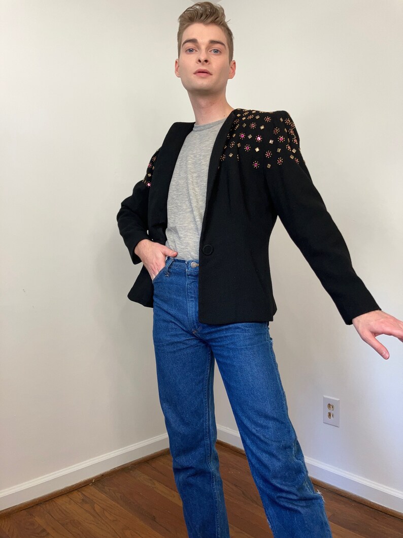 May include: A black blazer with a gray t-shirt underneath. The blazer has a unique design with pink and gold embellishments on the shoulders.