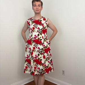 Peut inclure: Robe blanche sans manches à motif floral de roses rouges. La robe présente une encolure ronde et des poches. Les roses sont d'un rouge vif avec des feuilles vertes et des accents roses. La robe est de longueur genou et en tissu texturé.