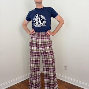 Könnte beinhalten: Ein dunkelblaues Kurzarm-T-Shirt mit einem weißen Grafikdruck von Bergen und dem Text RIO DE JANEIRO, kombiniert mit einer hoch taillierten Hose mit weitem Bein und einem Karomuster in den Farben Violett, Grün, Weiß und Schwarz.