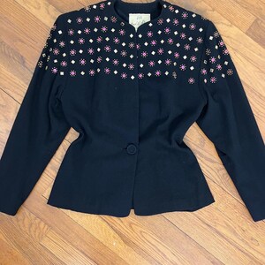 May include: A black blazer with a fitted silhouette and long sleeves. The blazer is adorned with a pattern of pink and gold embellishments across the shoulders and upper chest.