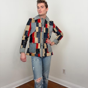 May include: A patchwork jacket with a grey collar, featuring a geometric pattern of red, black, tan, and blue squares. The jacket has long sleeves and a button-up front. The person is wearing light blue jeans with distressed details.