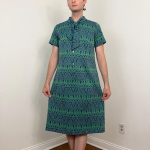 Può includere: Un abito vintage al ginocchio con un vivace motivo a zig-zag nelle tonalità del verde, blu e viola. L'abito presenta maniche corte, un'abbottonatura frontale e uno scollo a balze. L'abito è realizzato in un tessuto strutturato.