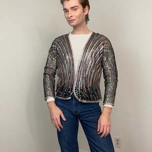 May include: A sequined jacket with vertical stripes in various colors, including silver, red, and blue. The jacket is worn over a white long-sleeved shirt and paired with blue jeans.