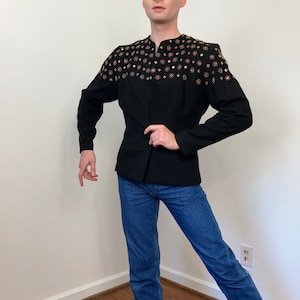May include: A black blazer with a stand-up collar and a row of gold and pink rhinestones across the shoulders. The blazer is worn over a pair of blue jeans.