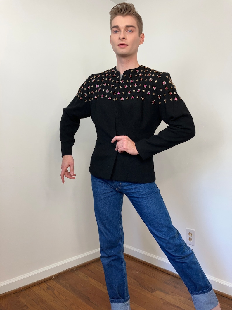May include: A black blazer with a stand-up collar and a row of gold and pink rhinestones across the shoulders. The blazer is worn over a pair of blue jeans.