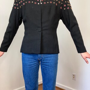 May include: A black blazer with a single button closure and a unique design featuring gold and pink rhinestones on the shoulders and collar.