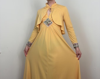 May include: A floor-length yellow gown featuring a matching cropped jacket with long sleeves. The dress has a high neckline with a tie closure and a diamond-shaped floral patterned inset at the chest. The sleeve cuffs also feature matching floral trim.