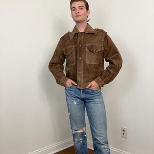 May include: A brown suede jacket with a collared neckline, button closures, and two chest pockets. The jacket is paired with blue jeans with distressed details and a rolled hem. The model is standing in a corner.